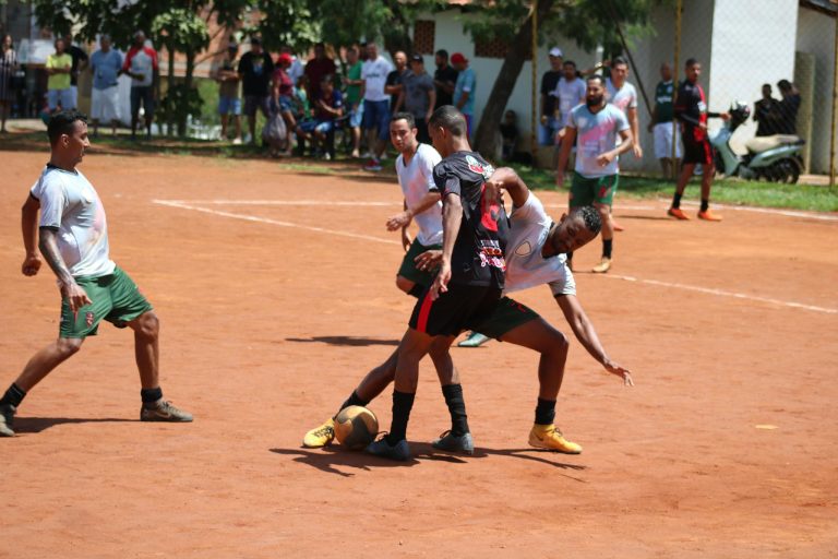 Players engage in a competitive soccer match on an outdoor dirt field with a crowd watching.