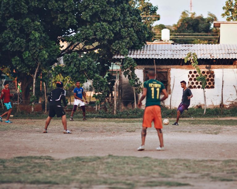 Teenagers play soccer outdoors on a dirt field in a casual urban setting.